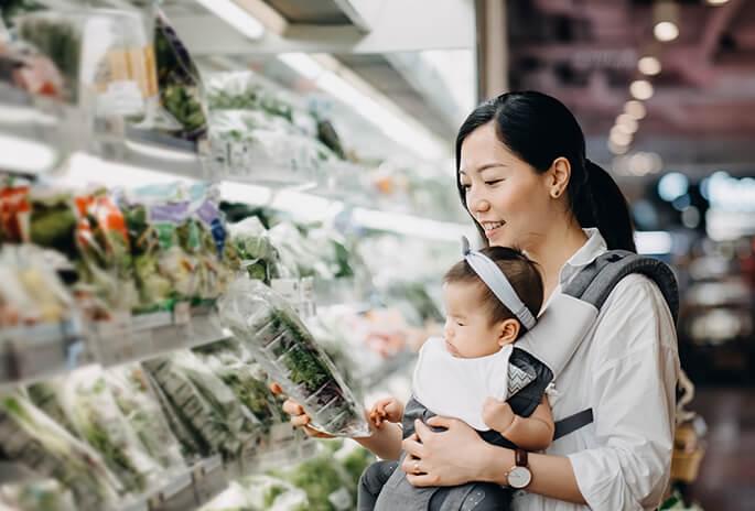 Woman with a baby in a carrier shopping for packaged salad